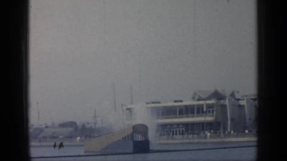 Distant fairgrounds at Flushing Meadows, with a pavilion, ramp, railings, and tiny visitor figures near the entrance.