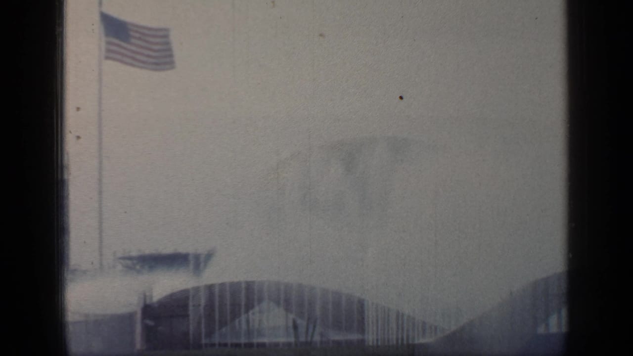 American flag flies over a hazy Flushing Meadows World's Fair scene with arched buildings and water spray below.