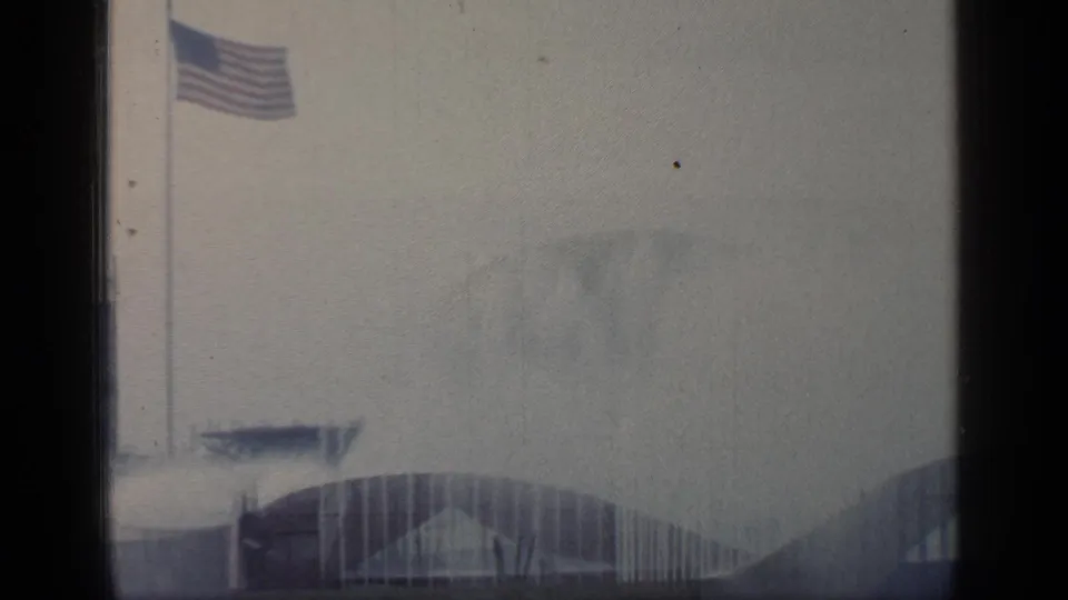 American flag flies over a hazy Flushing Meadows World's Fair scene with arched buildings and water spray below.