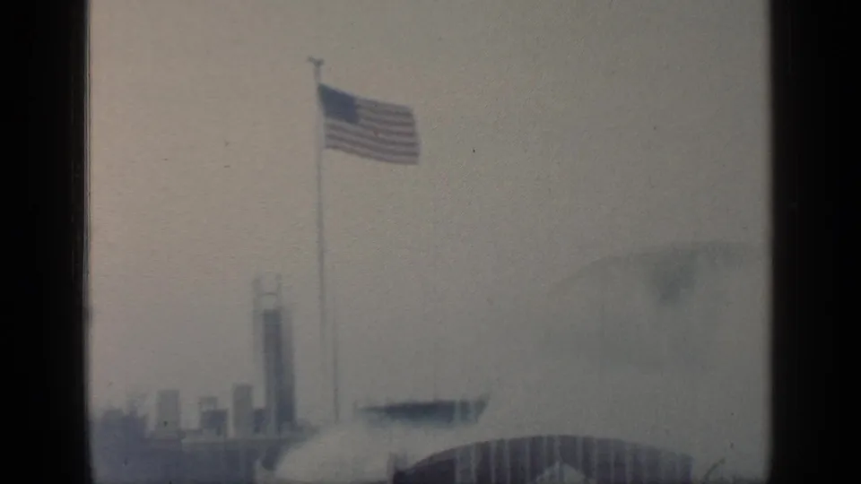 Visitor-shot 1964 Flushing Meadows scene with an American flag, slim towers, misty water feature, and arched fair architecture.