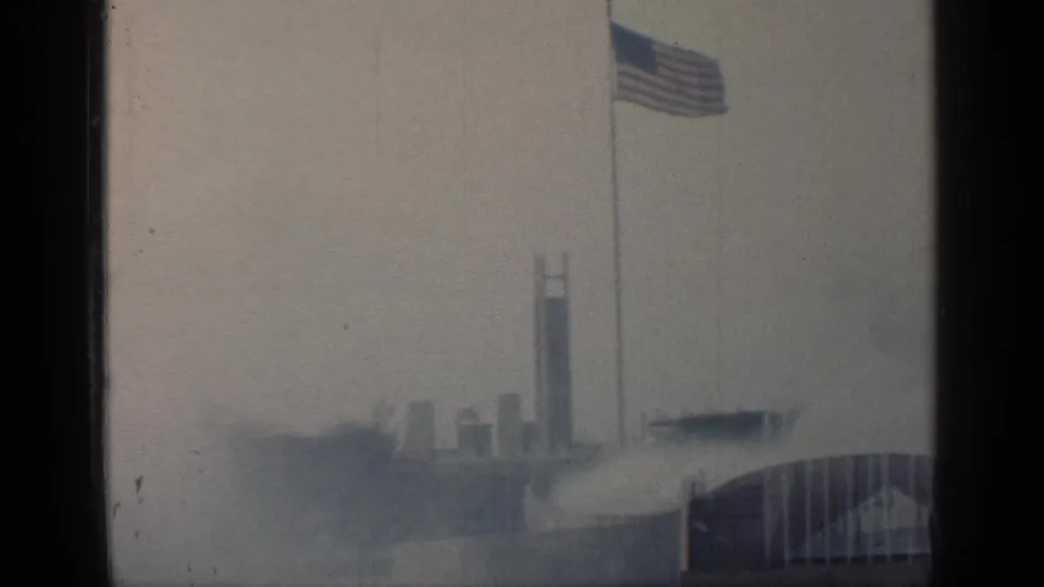 Hazy 1964 World's Fair view in Flushing Meadows with an American flag, vertical towers, water spray, and arched structure at the edge.
