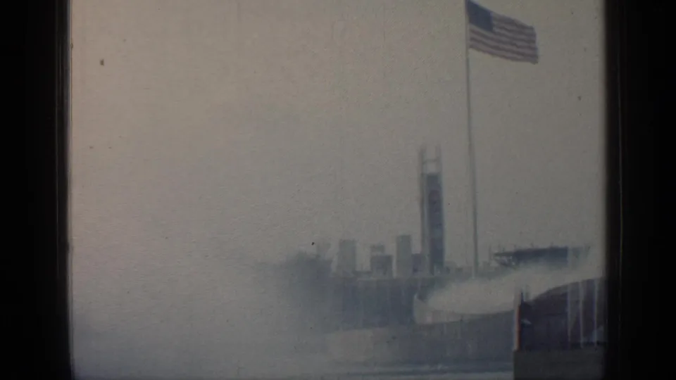 1964 Flushing Meadows home-movie frame with an American flag flying above distant World's Fair structures under an overcast sky.