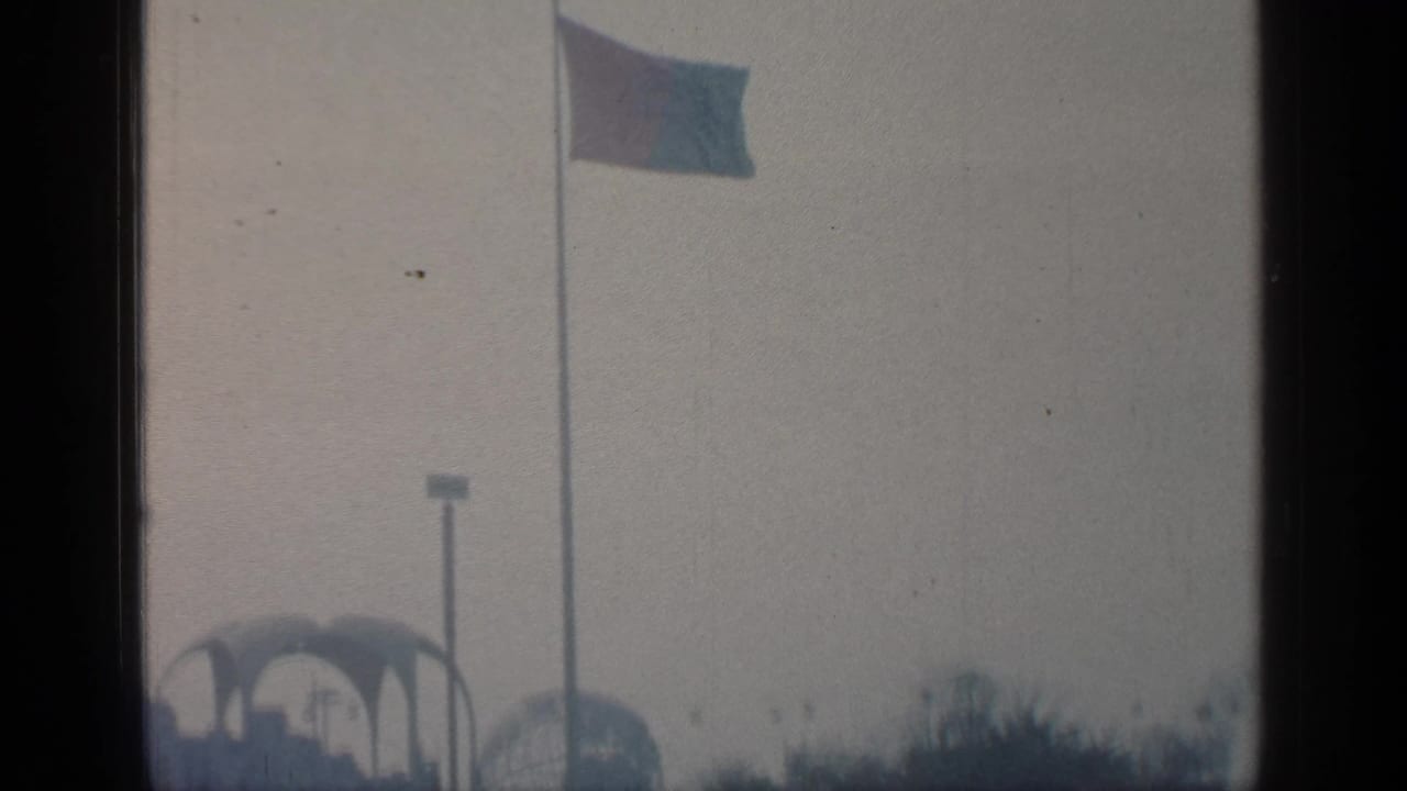 Distant fairground structures beneath a waving flag in a hazy sky.