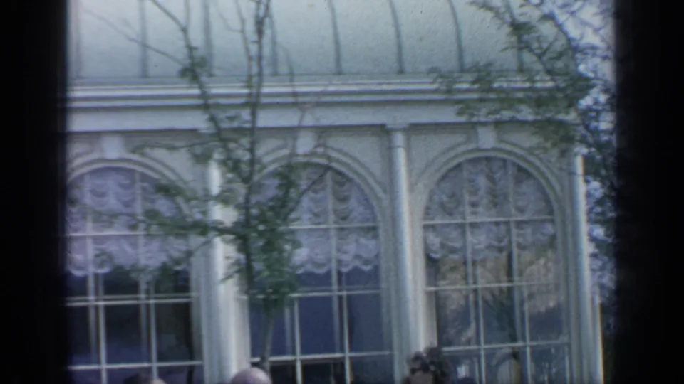 1964 Flushing Meadows view of tall arched windows, white columns, lace-like curtains, and trees from a handheld 8mm frame.