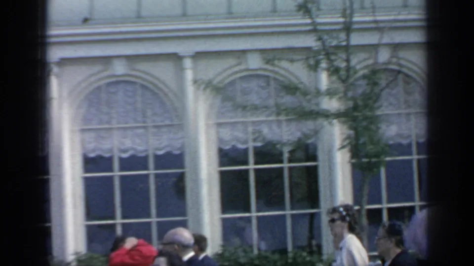 1964 Flushing Meadows visitors walk past arched windows, white columns, shrubs, and trees in a home-movie fairground view.