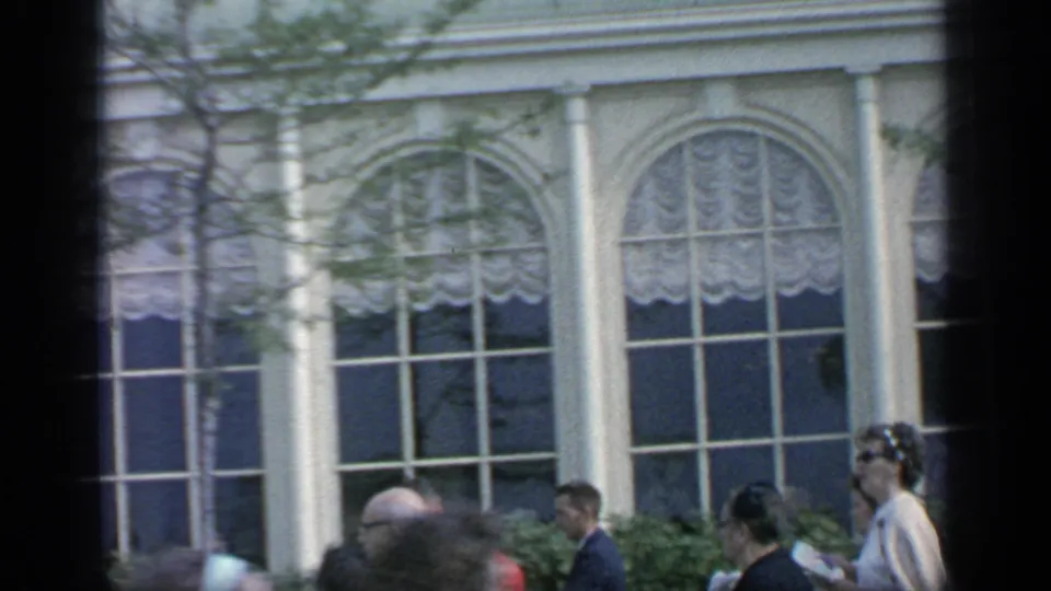 1964 Flushing Meadows crowd detail with suited visitors, arched windows, white columns, shrubs, and a handheld 8mm border.