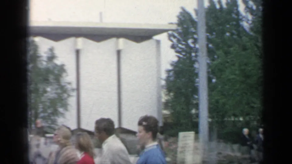 Visitors in 1960s clothing move through the World's Fair grounds beside a white pavilion structure and landscaped trees.