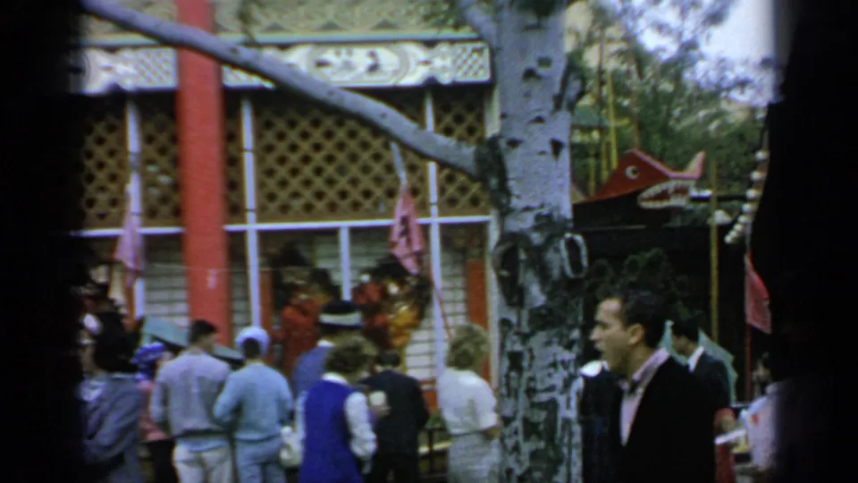 1964 World's Fair visitors cluster near a decorated pavilion, with red columns, patterned trim, and a birch tree in the foreground.