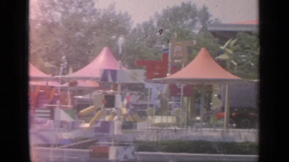 1964 Flushing Meadows fairground view with peaked tents, colorful ride structures, trees, and faded 8mm exposure.