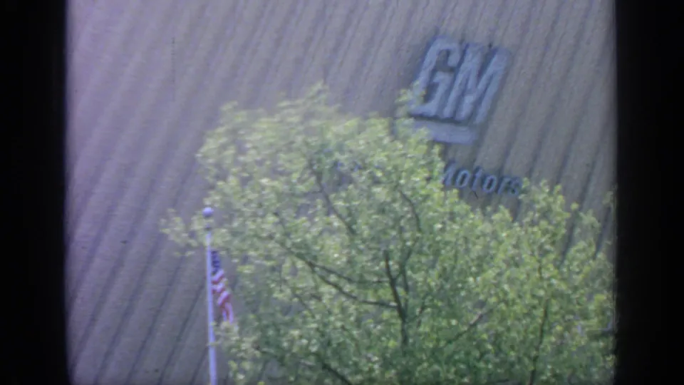 1964 Flushing Meadows frame showing the General Motors facade, an American flag, and leafy trees partly obscuring the building.