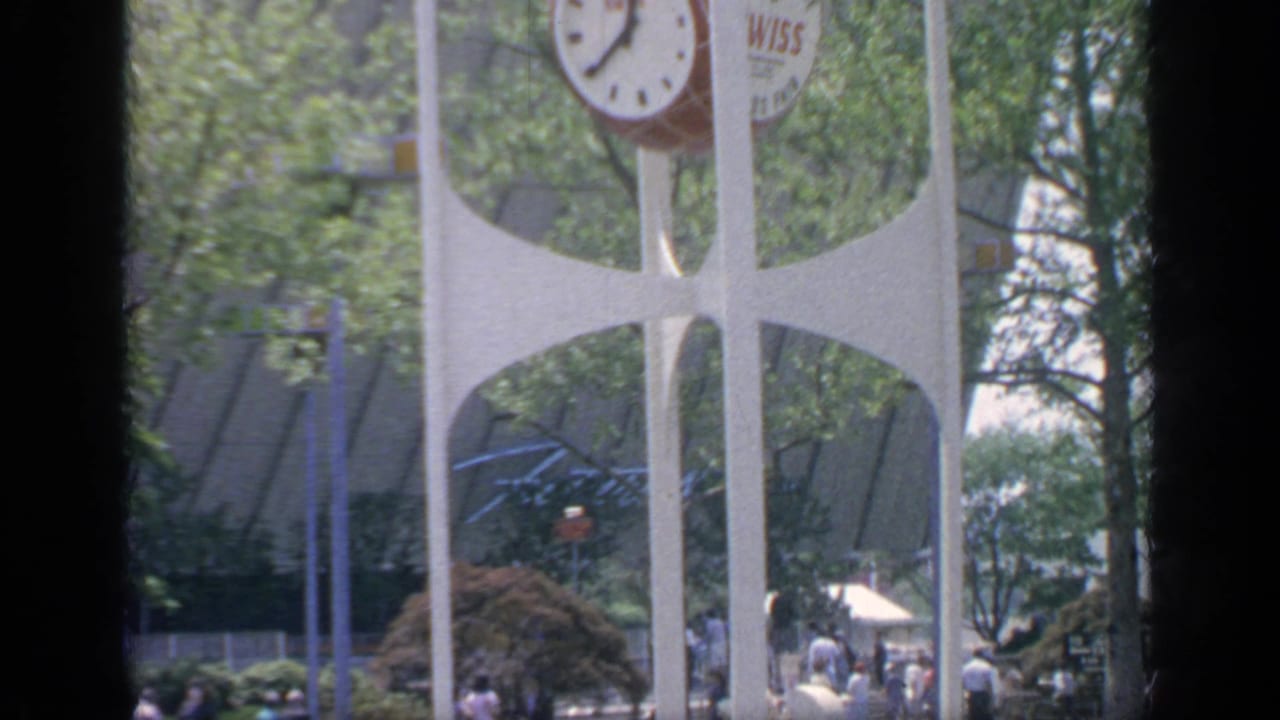 Visitor-shot 1964 World's Fair view of the Wiss clock tower, trees, and small figures moving through the grounds.