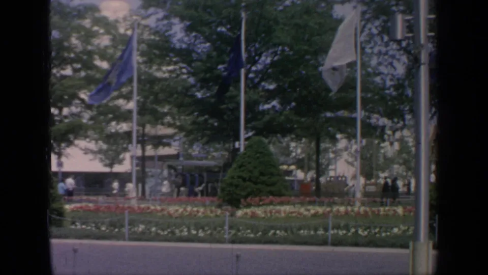 1964 World's Fair grounds with flagpoles, landscaped flower beds, trees, distant visitors, and exhibition buildings.
