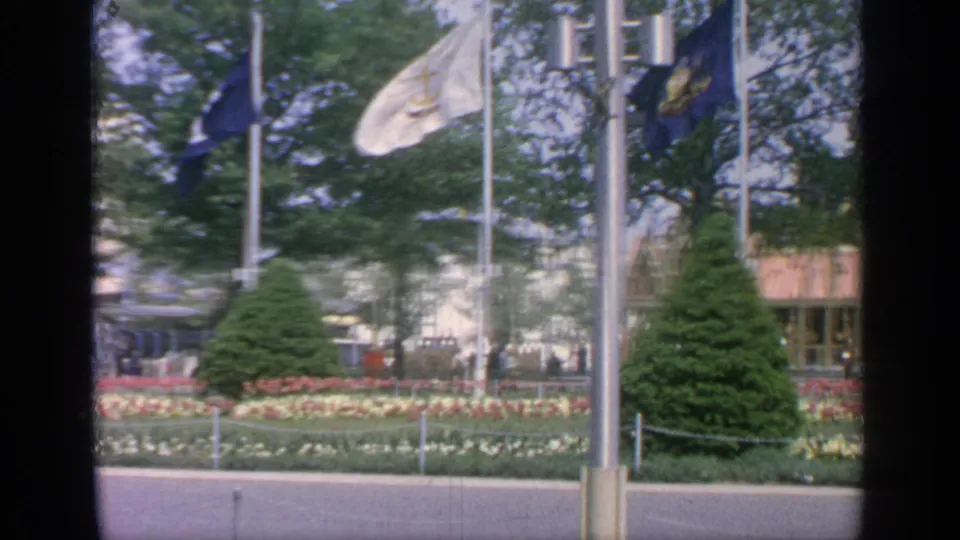 Flags flutter over manicured gardens at the Flushing Meadows fairgrounds, with trees, buildings, and small figures in view.