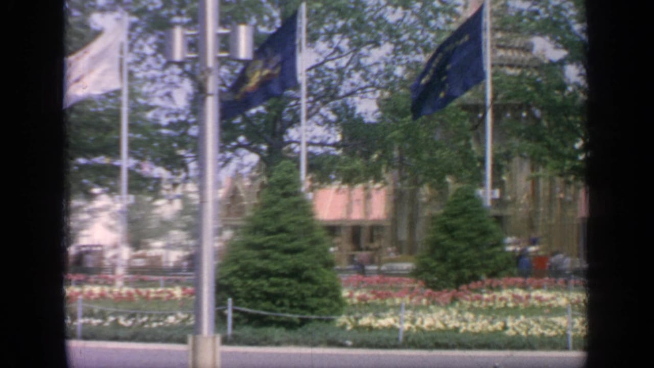 Flags on tall poles stand over flower beds and trees beside a fairground building.