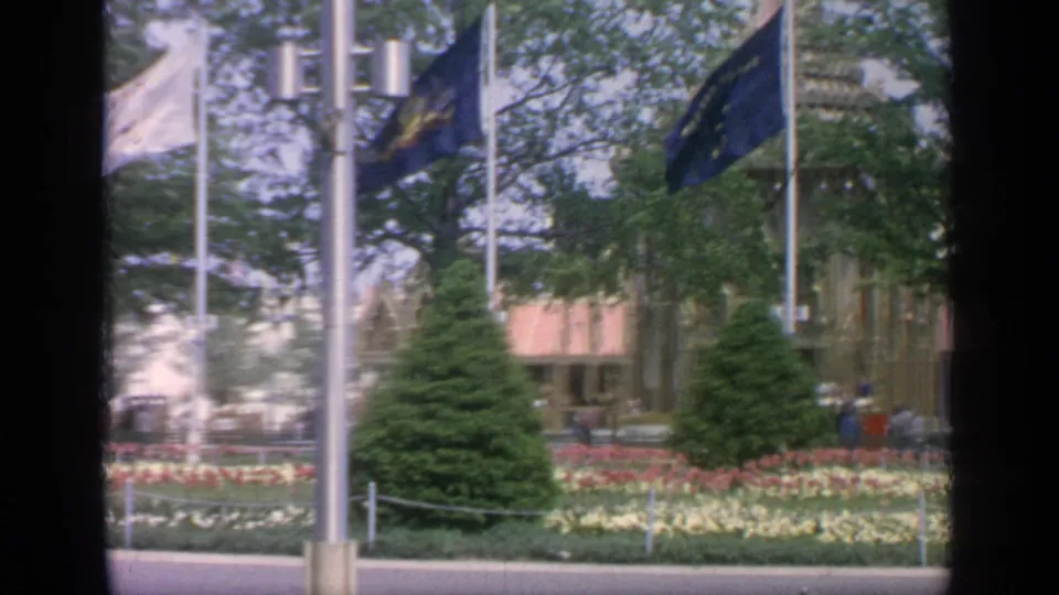 1964 Flushing Meadows view of blue and pale flags above formal flowers, trees and a fairground structure with visitors in the distance.