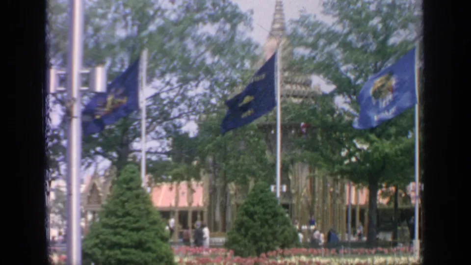 Visitor-shot 1964 Flushing Meadows footage frames blue flags, formal planting beds, evergreens and a decorative fairground building.