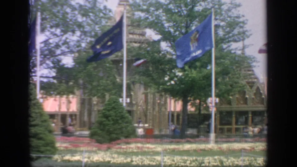 1964 Flushing Meadows fairground frame with blue flags, planted beds, trees, evergreens and distant visitors near a building entrance.