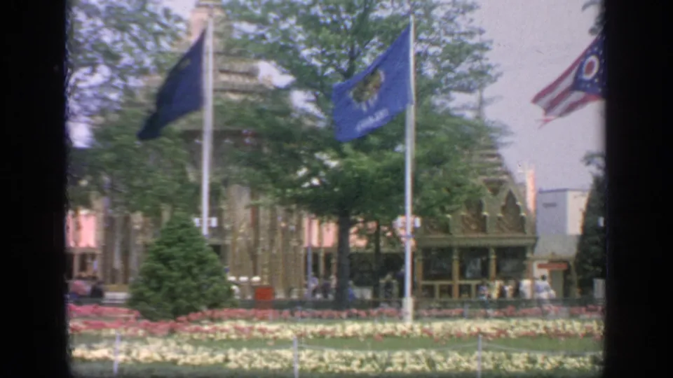 1964 Flushing Meadows fairground view with waving flags, patterned flower beds, shrubs, trees, and pavilion structures beyond.