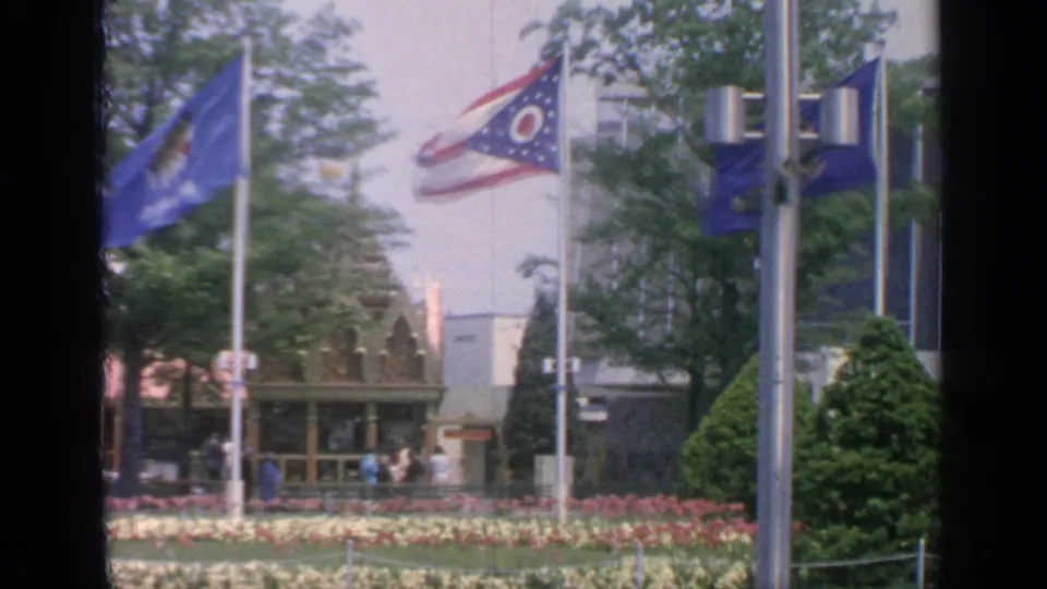 Visitor-shot 1964 fairground scene with blue flag, Ohio flag, flowers, trimmed greenery, trees, and pavilion exteriors.