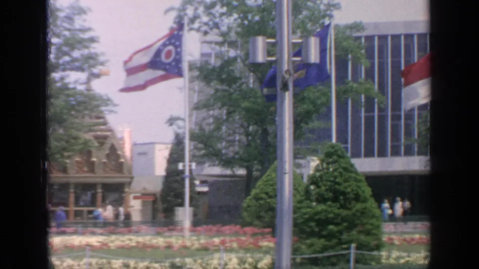 1964 Flushing Meadows view with Ohio flag, fair banners, flower beds, a modern pavilion facade, trees, and distant visitors.