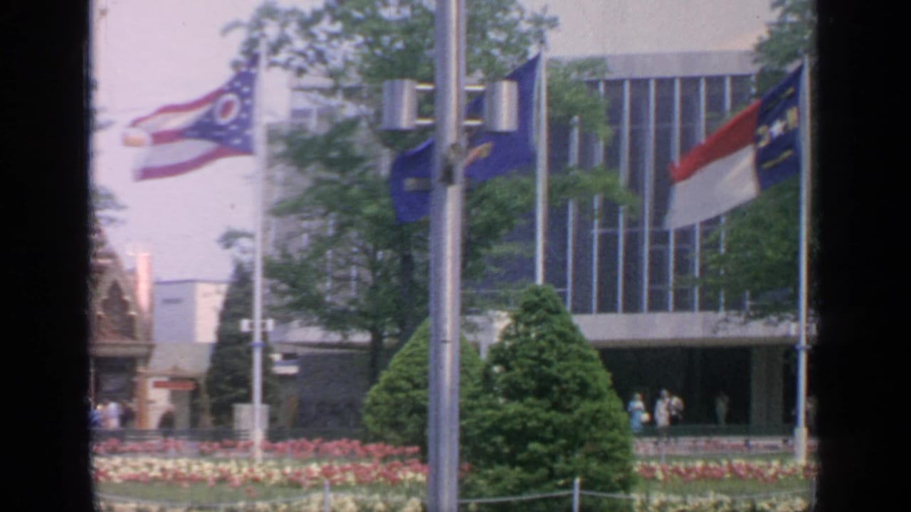 1964 Flushing Meadows fairgrounds view with flags, tulip beds, trimmed trees, a modernist building, and small visitors near the entrance.