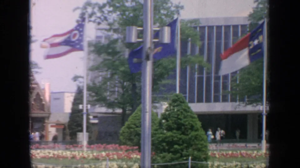1964 Flushing Meadows fairgrounds view with flags, tulip beds, trimmed trees, a modernist building, and small visitors near the entrance.