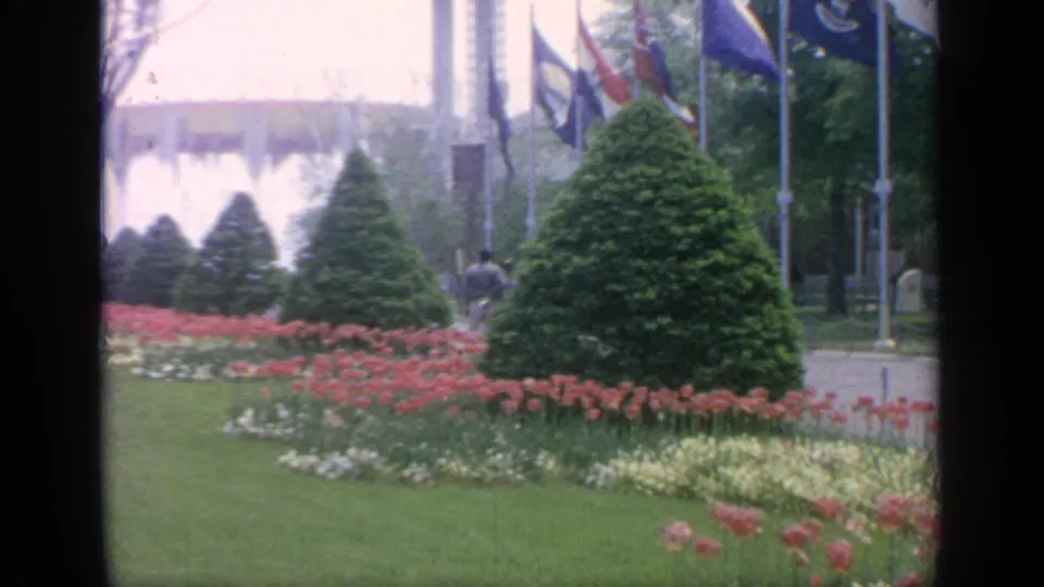 Flushing Meadows World's Fair landscaping with fountain spray behind flags, flower beds, clipped evergreens, and open lawn.