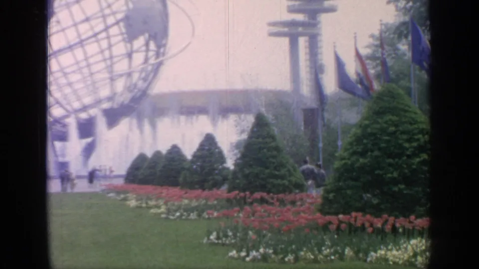 The Unisphere appears at the edge of the frame above fountain spray, flags, flower beds, trimmed evergreens, and distant fair visitors.