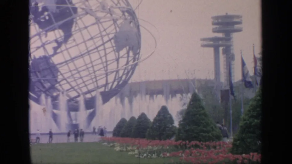 Wide fairgrounds view with the Unisphere, fountain spray, flower beds, trimmed evergreens, flags, and small visitor figures in the distance.