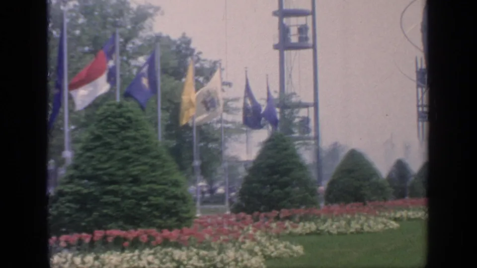 1964 World's Fair garden view with flags, an observation tower, fountains, trimmed shrubs, flowerbeds, and green lawn at Flushing Meadows.