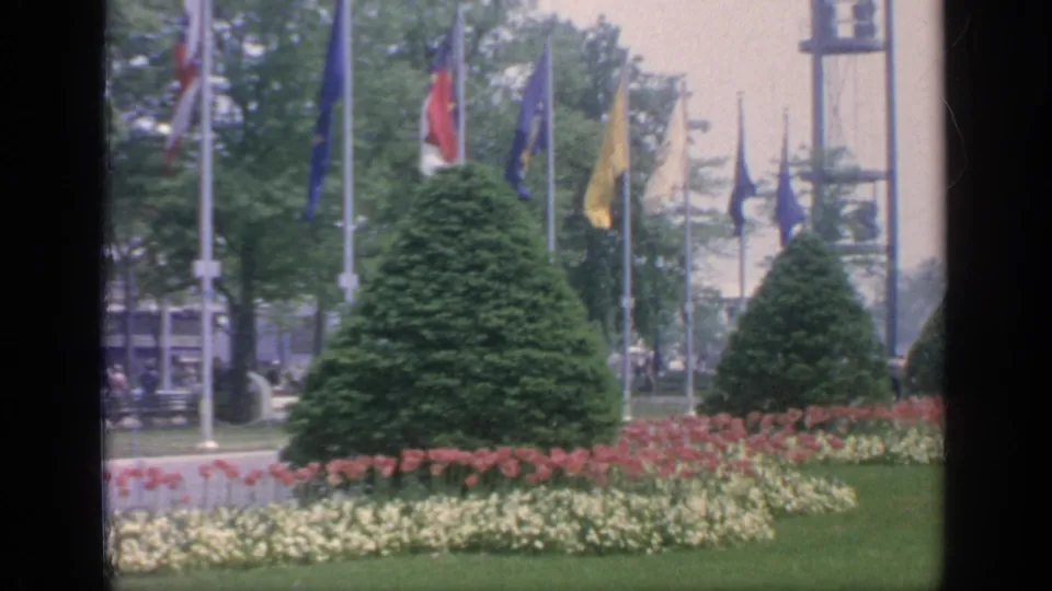 Flushing Meadows fairground landscaping with waving flags, a large conical shrub, flowerbeds, lawn, trees, and an observation tower in the background.
