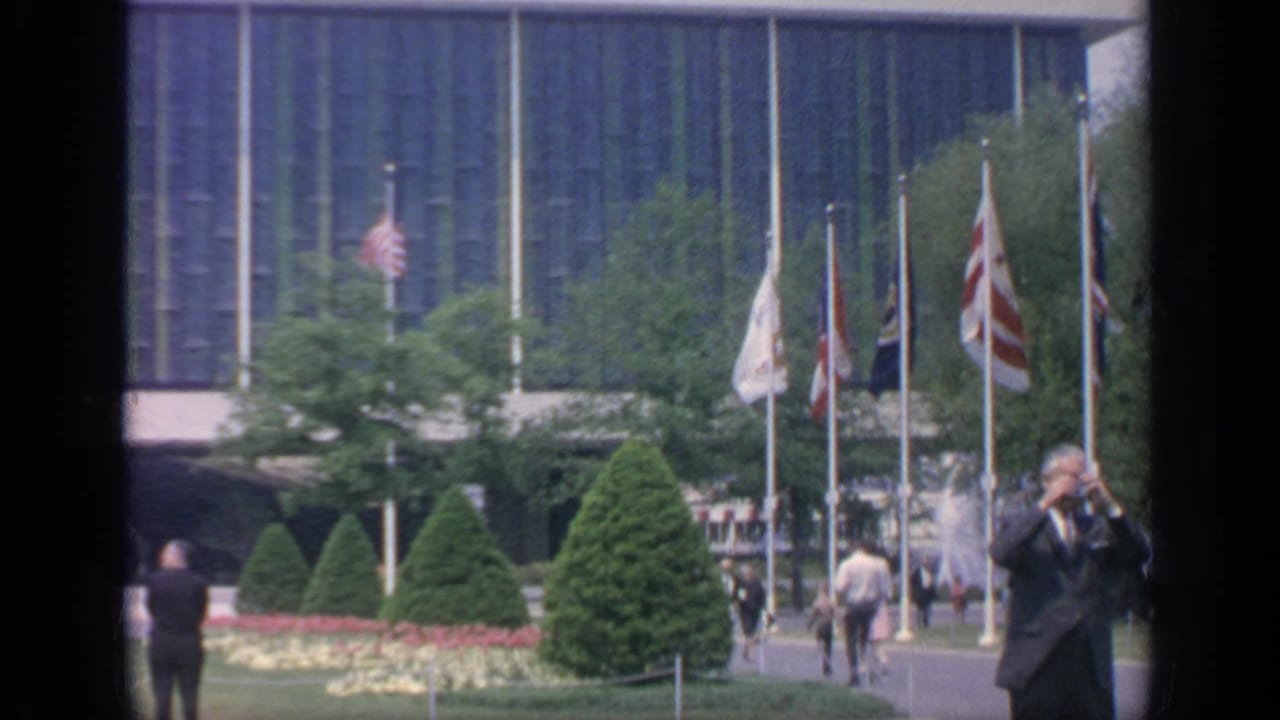 Visitors walk across a landscaped courtyard with flags, shrubs, flowerbeds, and a glass-fronted building behind them.