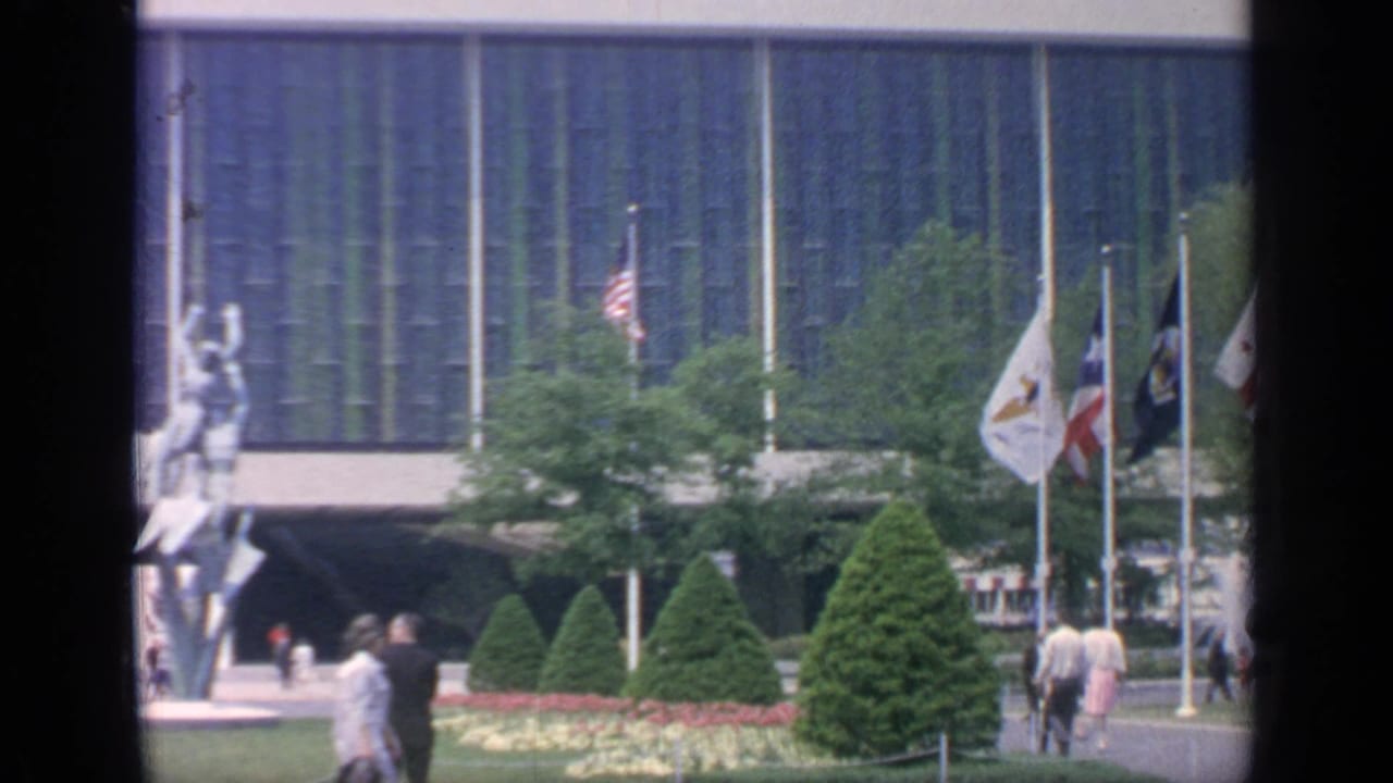 A courtyard with flags, pedestrians, conical shrubs, flowerbeds, and a tall outdoor sculpture near a glass building.