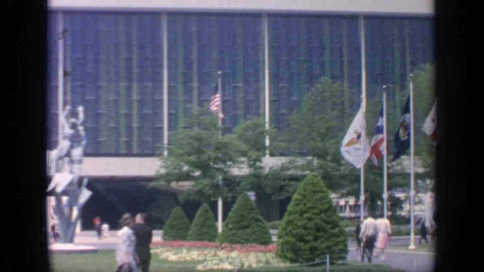 1964 Flushing Meadows frame with visitors walking past flags, manicured plantings, and a prominent sculpture outside a modern building.
