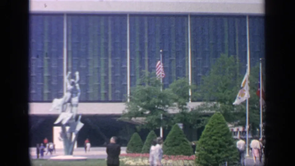 1964 Flushing Meadows footage frame showing a sculpture, flags, visitors, clipped greenery, and the blue-green facade of a fairground building.