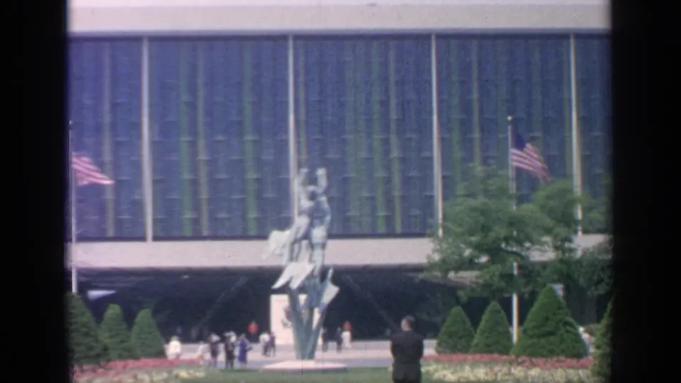 1964 Flushing Meadows frame with a tall outdoor sculpture, American flag, visitors, trees, and modern glass architecture in view.
