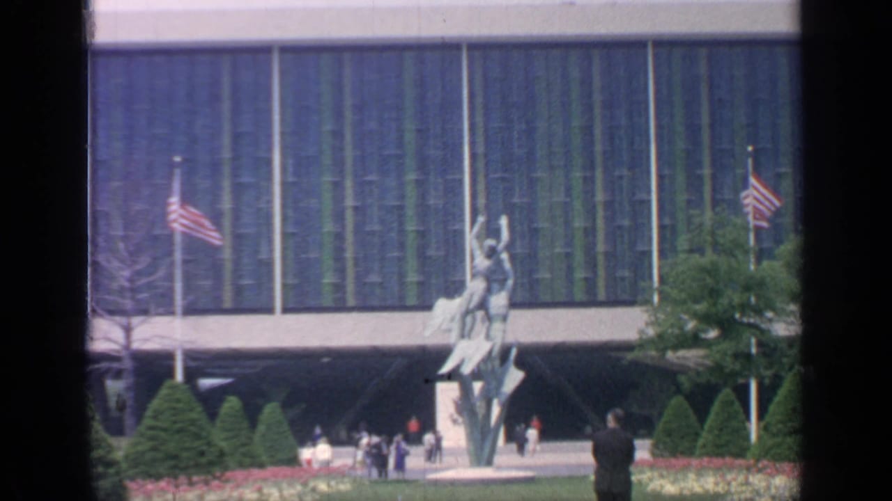 Visitors pass a modernist building with flags, sculpture, evergreens, and flower beds.