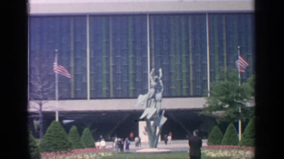 1964 Flushing Meadows visitor view of a blue-green modernist facade, American flags, a tall sculpture, trimmed trees, and people in the courtyard.
