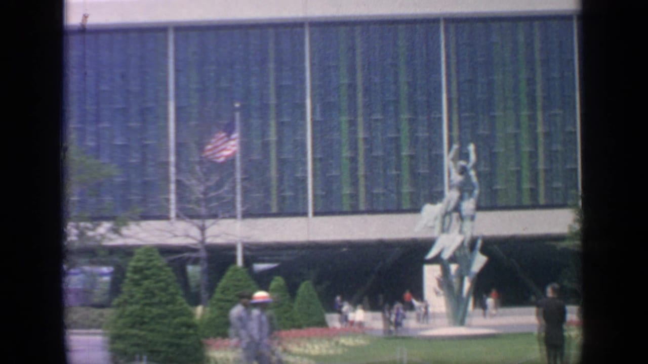A sculpture stands near flags and visitors in front of a blue-green facade.