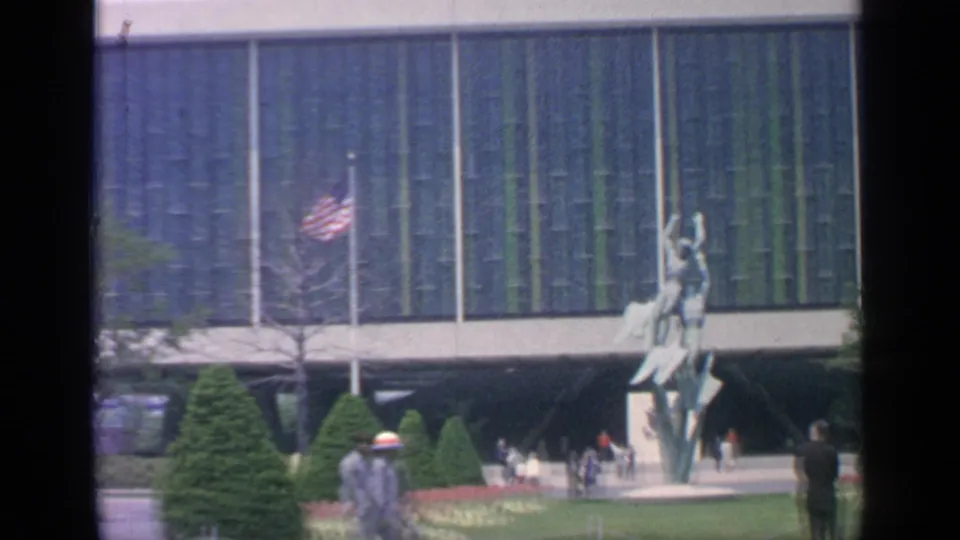 At the 1964 Flushing Meadows fairgrounds, visitors walk near a tall sculpture, flagpoles, clipped landscaping, and a large striped glass building.