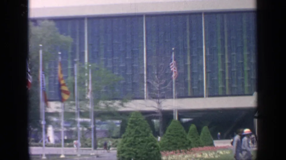 A visitor-shot 1964 Flushing Meadows view shows multiple flagpoles, manicured evergreens, people on walkways, and the blue-green facade behind them.