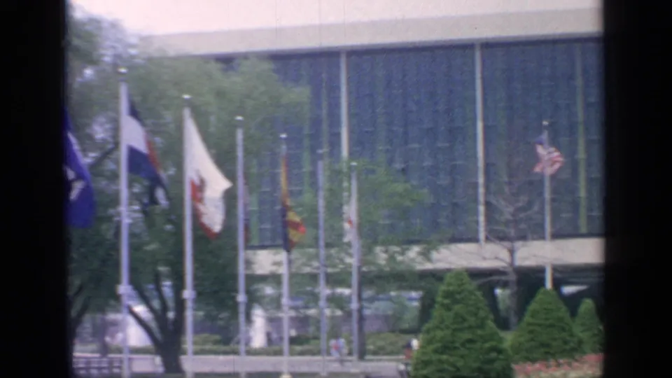 The 1964 Flushing Meadows courtyard view includes international flags, a large striped glass building, trees, flower beds, and visitors walking through the grounds.