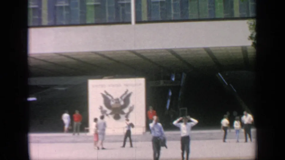Flushing Meadows fairground plaza with scattered visitors, a large eagle-emblem panel, and a shadowed modern structure.