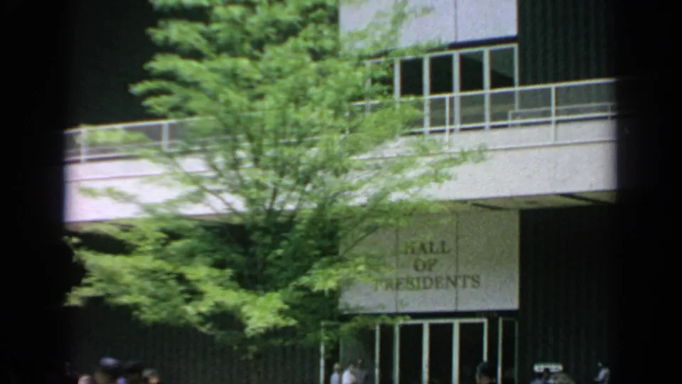 1964 World's Fair Hall of Residents entrance partly framed by trees, with signage, glass doors, and distant visitors.