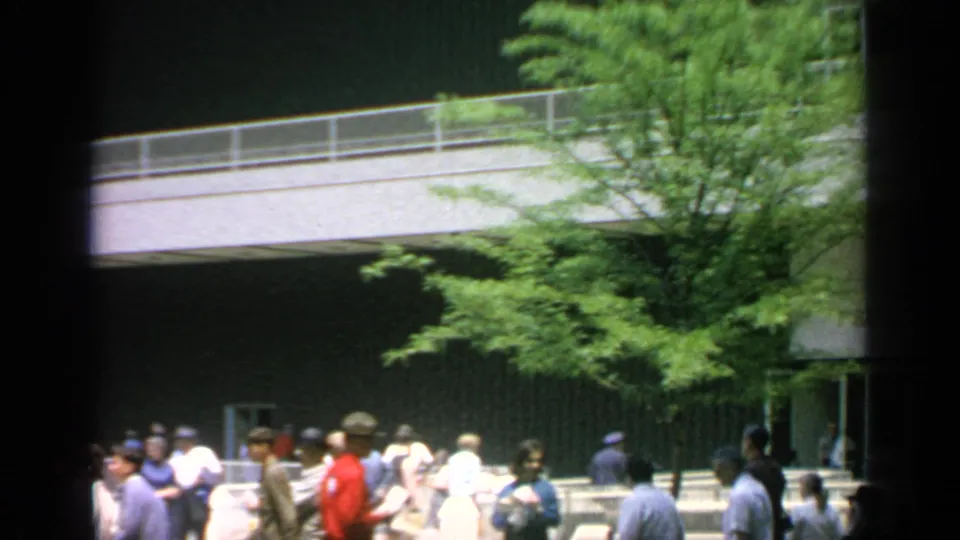 Visitors in 1960s clothing walk outside the Hall of Residents at Flushing Meadows, with trees and modern facade behind them.