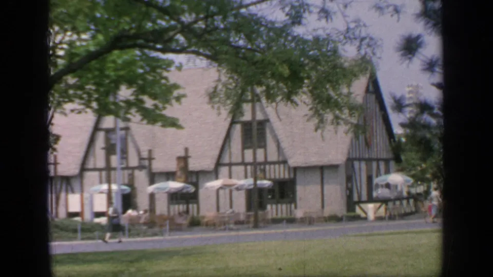1964 Flushing Meadows fairground scene with a Tudor-style building, patio umbrellas, and distant visitors on the path.