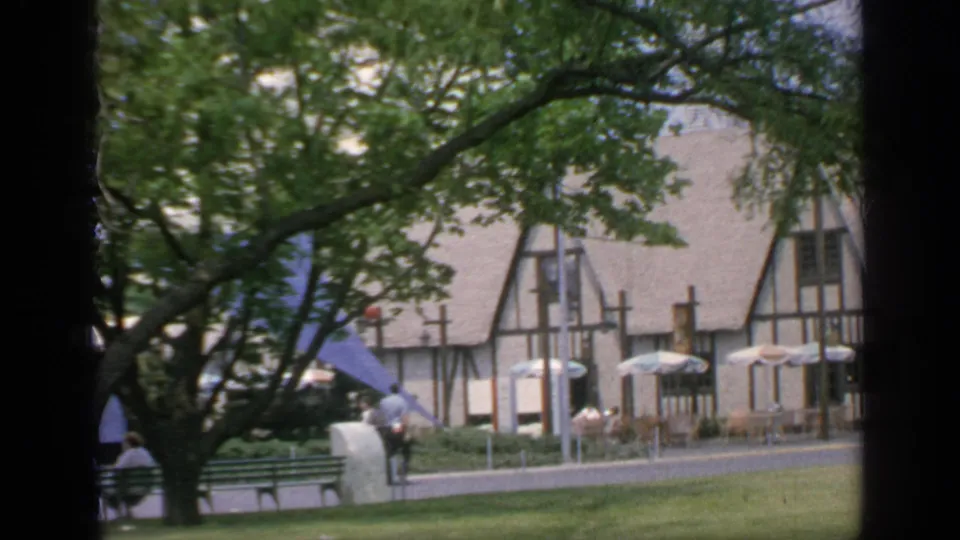 1964 Flushing Meadows view through a large tree toward the Tudor-style building, umbrellas, benches, and fair visitors.