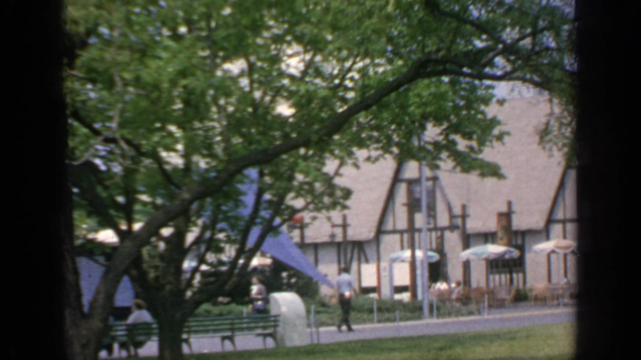 Tree-framed Tudor-style building with benches, umbrellas, and visitors on a walkway.