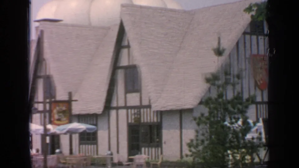 Home-movie view of the 1964 Flushing Meadows Tudor-style building with outdoor umbrellas, seating, and a red shield sign.
