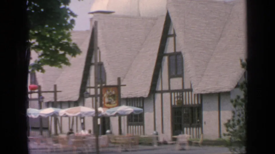 1964 Flushing Meadows frame with a Tudor-style building, pale steep roof, patio umbrellas, chairs, trees, and a red shield emblem.